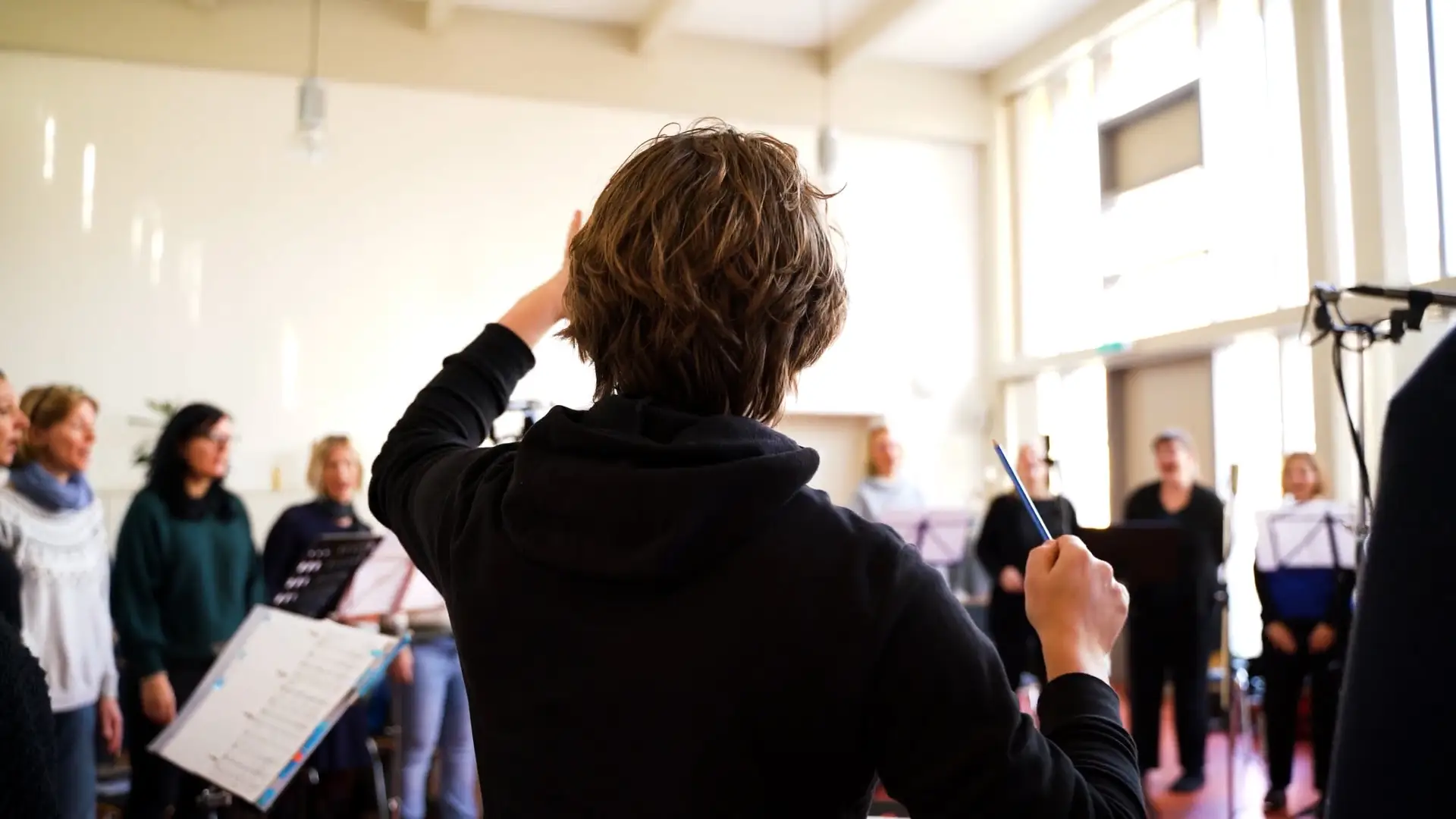 Ein Foto hinter einem Dirigenten. Unscharf im Hintergrund sieht man den Chor. Es sind Notenständer, sowie Mikrofone verteilt und aufgestellt.