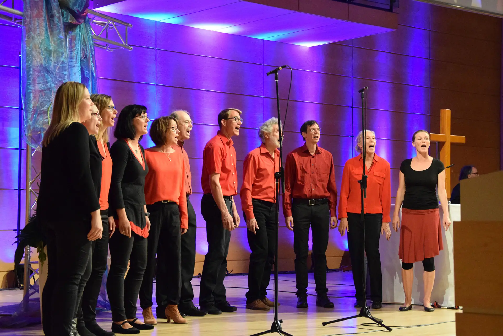 Eine Gruppe des Jazzchors inTune singt auf einer Bühne in einem modernen Saal mit violetter Beleuchtung.